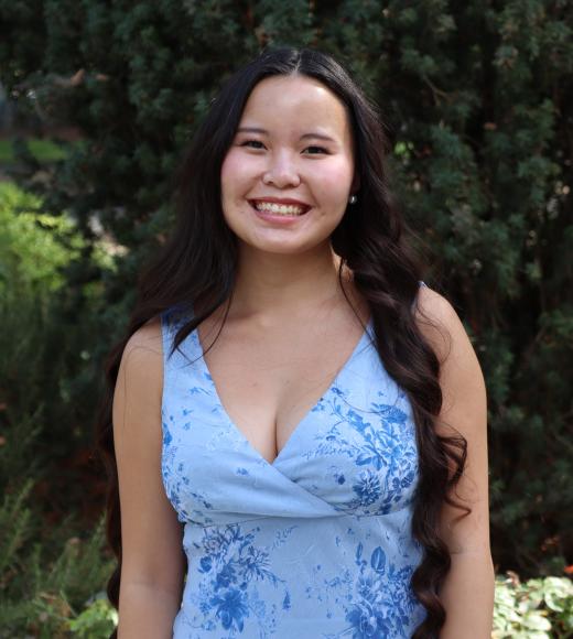 Profile picture of Aneka wearing a light blue blouse and standing in front of a tall green tree.