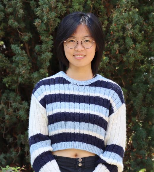 profile picture of Carly standing in front of a tall green tree, she is wearing glasses and a light blue, dark blue, and white striped shirt.