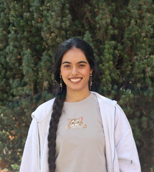 Photo of Jasmeet standing in front of tall green trees, she is wearing a light green shirt and white jacket. 