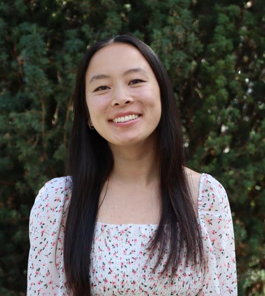 Profile picture of Kaley standing behind a tall green tree and wearing a white blouse
