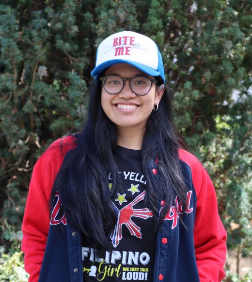 Profile picture of Lexi standing behind a tall green tree wearing a long sleeve red and black shirt and blue and white baseball cap