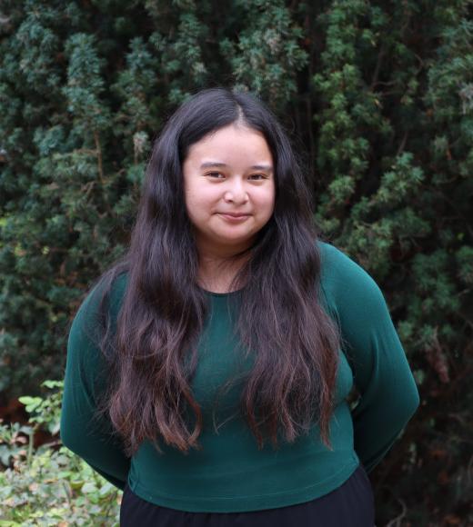 profile photo of Melissa standing in front of tall green trees. She is wearing a dark green sweater and black pants