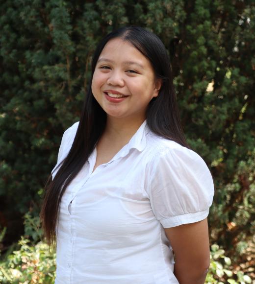 Profile picture of River standing in front of a tall green tree. She is wearing a white shirt.