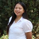 Profile picture of River standing in front of a tall green tree. She is wearing a white shirt.